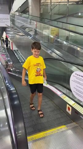 A young boy wearing a yellow t-shirt with a cartoon character on it and black shorts, brown sandals, stands on an escalator in a well-lit indoor environment. He holds a small object, possibly a toy, in his right hand. The escalator has a dark gray tread and a yellow safety line. He smiles at the camera and then turns to his left, looking down the escalator. The background shows another escalator going in the opposite direction, with people on it, and glass railings. There are signs in the background, one with Vietnamese text. The boy then turns back to the camera.
