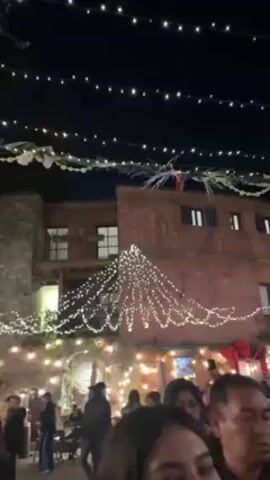 The video captures an outdoor scene at night, likely a public space or courtyard, adorned with festive lighting and Christmas decorations. Strings of lights are hung overhead, creating a canopy of illumination, and more lights drape from the roofs of the buildings. The buildings themselves are of varying heights and constructed from what appears to be brick or stone.

People are present in the space, moving about and interacting with each other, although their activities are obscured by the crowd. In the foreground, a person is visible, looking toward the camera, possibly filming the scene. The overall atmosphere appears festive and lively.