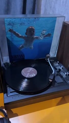 A Nirvana album cover featuring a baby swimming underwater with a dollar bill is shown behind a spinning vinyl record on a turntable. The turntable is black with silver accents and a black tonearm. The background is dark with a blue tint. The scene is lit by a warm yellow light from below, casting shadows on the turntable.