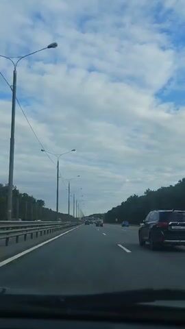 A first-person perspective shot captures a view from inside a vehicle driving on a multi-lane highway. The sky is predominantly blue with scattered white clouds. Tall lampposts line the left side of the highway, which is separated from the adjacent area by a metal barrier. Trees and foliage form a dense line along the right side of the road. Several vehicles, including cars and what appears to be a taxi, are visible on the highway, traveling in the same direction. The road surface is asphalt, marked with white lines.