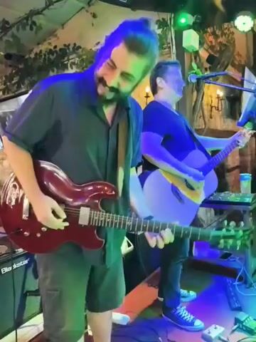 The video shows a band performing in what appears to be an outdoor restaurant or venue. Two men are visible on stage, one playing an electric guitar and the other an acoustic guitar and singing. The electric guitarist is in the foreground, wearing a dark blue shirt and dark shorts. The acoustic guitarist is in the background, wearing a black t-shirt and dark pants. Both are smiling and engaged in the performance. The stage is lit with colorful lights, including pink and purple. In the background, patrons are seated at tables, some of whom are looking towards the stage. The venue is decorated with lights and foliage. The overall atmosphere seems lively and festive.