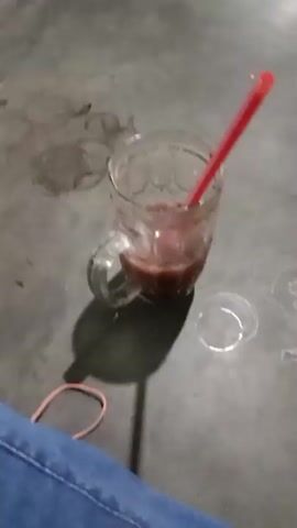 The video pans across a dimly lit, somewhat rustic indoor space. In the foreground, a glass with a reddish-brown liquid and a red straw sits on a concrete floor. Further back, several large plastic water jugs are visible, along with a green bucket and a red broom. In the mid-ground, two men are seated at a wooden table, with one man facing the camera and another turned away. Behind them, a television screen displays a bright, colorful image. The walls and ceiling appear to be constructed from corrugated metal and rough wooden beams, suggesting a simple or utilitarian structure. The overall impression is of an informal gathering space, possibly a small shop or eatery.