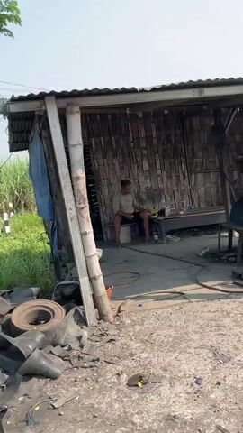 A man sits on a stool inside a small, open-air structure with a corrugated metal roof and bamboo walls. The structure appears to be some kind of workshop or shed. In front of the structure, there are discarded tires and other debris on the ground. To the left of the structure, there is tall grass and vegetation. The lighting suggests it is daytime, possibly late morning or afternoon, with clear skies.