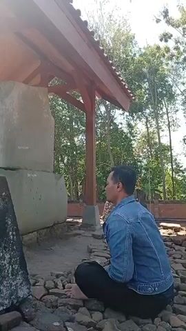 A man wearing a denim jacket and black pants kneels on the ground in front of a large stone monument. The monument is partially covered by a wooden shelter with a terracotta roof. The ground around the monument is covered in stones and dirt. Trees and foliage are visible in the background. The man appears to be looking at the monument.