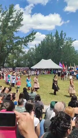 The video shows an outdoor event with a crowd of people watching a procession. In the center of the frame, several individuals in traditional Indigenous regalia are walking across a grassy field. They are wearing colorful dresses and headpieces. Behind them, a large crowd of spectators is gathered, some holding up phones to record the event. A white tent is visible in the background, along with trees and a blue sky with white clouds. The event appears to be a cultural gathering or celebration.