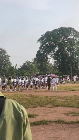 In this video, a large group of people, likely young adults, are gathered outdoors in a field. They are dressed in matching uniforms: white t-shirts or tops and green shorts, with some wearing green caps. The individuals appear to be participating in some form of organized activity, possibly a parade or training exercise, as they are in formation and moving in unison.

The scene is set in a rural or semi-rural area. The ground is covered with dry, patchy grass and dirt. In the background, there are numerous trees, including a very large, mature tree dominating the right side of the frame. Some buildings and structures are visible beyond the trees, suggesting the presence of a settlement or institution. The sky is bright and appears to be clear, indicating a daytime setting. The overall mood seems to be one of order and discipline, given the uniform attire and synchronized movements. There are a few individuals in different attire, including one person in a military-style uniform, who might be an instructor or supervisor.

While no specific text is clearly legible in the video, the uniforms suggest a group with a shared purpose, possibly a youth corps or a similar program common in Nigeria, where the location context is Ise- Ekiti. The activity appears to be taking place in the morning or early afternoon, given the sunlight. The weather seems fair, suitable for outdoor activities.