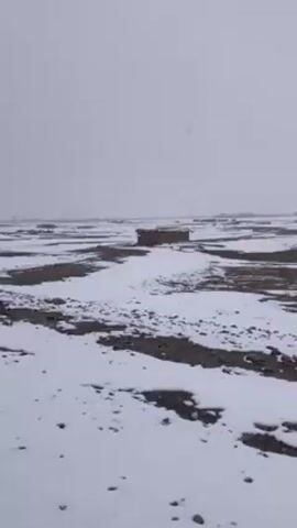 The video depicts a desolate, flat landscape partially covered in snow. The ground is a mix of dark earth and white snow patches, creating a stark contrast. In the distance, there are several indistinct, low-lying structures scattered across the terrain. The sky is overcast and a pale gray, suggesting a snowy or wintery day. No people or animals are visible in the scene. The overall impression is one of emptiness and cold.