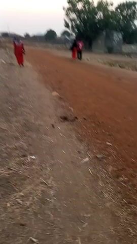 The video shows a dirt road with a couple walking away in the distance. The road is reddish brown and the sides are dry with some scattered debris. There's a small sign on the right side of the road and some trees in the background. The camera seems to be handheld and moves quickly, focusing mostly on the ground.