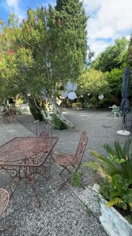 The video shows an outdoor patio area, likely at a restaurant or cafe, with several wrought iron tables and chairs arranged on a gravel surface. The patio is surrounded by trees and bushes, with hanging white lanterns visible in the branches of one tree. In the foreground, a square table with ornate metalwork is surrounded by matching chairs. A closed black umbrella stands next to a railing overlooking a body of water, possibly a lake or sea, under a partly cloudy sky. In the distance, buildings are visible along the shoreline. There are no people visible in the video.