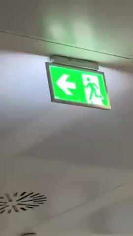 The video shows an illuminated green exit sign hanging from a white ceiling. The sign displays a white figure running towards a door with a white arrow pointing left.  An air vent is visible in the bottom left corner of the frame.  Audio indicates people are talking in the background, although the words are not distinct.