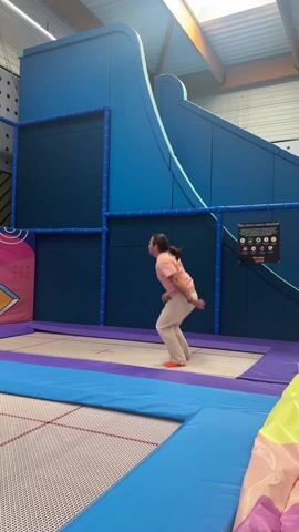 A person is jumping on a trampoline. They are wearing a pink shirt, white pants, and orange socks. The trampoline is surrounded by blue padding, and there is a large blue structure behind the trampoline. The background includes elements with multicolored and geometric designs. The scene appears to be inside a trampoline park, indicated by the "Gravity Jump" sign visible in the background. The person is mid-air, with their arms outstretched.