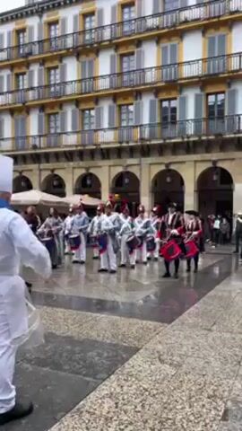 Here's a description of the video:

The video captures a vibrant and colorful scene in a European town square. It is likely a celebration, parade, or performance, as a group of musicians is playing drums while moving in unison. A number of drummers are attired in white chef uniforms including chef hats, some with blue neckerchiefs, and plastic rain gear. There are also several band members in more traditional red, white, and black military-style outfits with plumed hats. The drummers move from left to right. A man facing the camera in a chef's uniform appears to be leading the band.  The square is paved with geometric tiles, and a multi-story building with balconies and arched walkways surrounds the square. Patrons sit in an open cafe area under umbrellas. Overall, the scene is lively and full of energy.