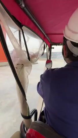 The video shows a person from behind, wearing a blue jumpsuit and a white hard hat, driving a small, open-air vehicle with a red canopy and clear plastic sides. The person is holding onto a steering wheel. The vehicle appears to be on a paved surface, possibly a construction site or industrial area, with stacks of grey materials on pallets visible to the left. In the background, there are buildings and structures. The camera is positioned inside the vehicle, looking forward and to the side, capturing the driver's perspective. The lighting suggests it is daytime.