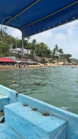 The video opens with a view from the stern of a light blue boat with a dark blue canopy, looking towards a sandy beach. The water is green with yellow buoys visible. On the beach, numerous people are present, alongside several buildings featuring red roofs and various umbrellas and tents. Behind the beach, a steep, green, tree-covered hillside rises, dotted with white buildings. At 0:02, a man wearing a blue t-shirt and patterned shorts enters the frame, standing in the back of the boat, facing away from the camera. He leans over the stern, appearing to interact with an engine. A red fuel tank is visible on the floor of the boat next to him. The boat slowly approaches the shore.