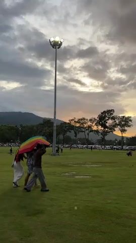 A group of three people walk across a grassy field under a multicolored umbrella. The sky is overcast with the sun setting in the distance, casting a warm glow through the clouds. A tall light pole with several floodlights stands in the foreground, illuminating the area. In the background, trees line the edge of the field, and a parking lot filled with cars is visible. Mountains form the horizon. The people are walking from left to right across the frame. One person in white pants walks on the far left, followed by a person in a patterned dress, and then a person in gray pants. The umbrella is held above them.