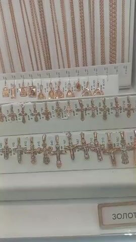 The video shows a display of gold jewelry in a store. The jewelry includes necklaces, pendants, and bracelets, with a particular emphasis on cross pendants. The items are neatly arranged in display cases, with price tags visible on some of the items. The lighting is bright, illuminating the gold jewelry. The overall impression is that of a well-stocked jewelry store.