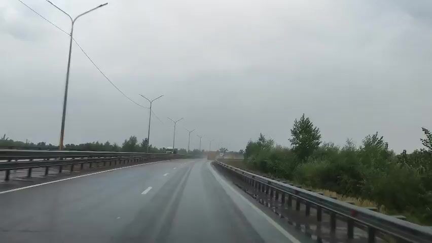 The video shows a first-person view of a car driving on a wet highway under a cloudy sky. The road is asphalt and appears to be in good condition with clear white lane markings. The car is in the right lane, and there are other vehicles visible in the distance, including a white truck on the left lane. On the right side of the road, there is a guardrail and dense green trees and bushes. On the left side, there is another guardrail and a few trees. Several lampposts line both sides of the highway. The overall impression is that of a normal drive on a highway during a cloudy or rainy day.