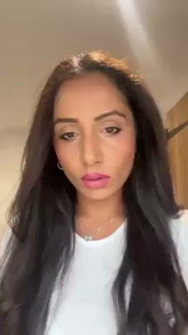 A woman with long dark hair and fair skin is applying makeup in a close-up video. She is wearing a white t-shirt, pink lipstick, and a silver necklace. She holds a makeup brush with a pink handle in her right hand and applies a pink substance to her right cheek. The background is a white wall and a brown door.