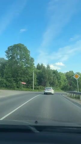 A driver's perspective shot shows a car driving on a winding road with lush green trees on both sides. The road appears to be a two-lane highway with a solid white line on the right shoulder and a dashed white line separating the lanes. There is a white sedan ahead of the camera car, and other vehicles can be seen in the distance. A blue sky with scattered clouds is visible overhead. The camera car passes a road sign indicating a 20-ton weight limit. The camera car then crosses a bridge. The setting appears to be a sunny day in a rural or suburban area.