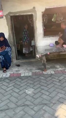 A woman wearing a blue hijab and a blue dress sits on a low, dark-colored stool to the left of a doorway, looking down at a mobile phone in her hands. She is sitting on a tiled walkway. To the right of the doorway, a man wearing a blue t-shirt and black shorts is crouched on a rustic wooden table that is positioned in front of a window opening. He is facing the window and appears to be looking or reaching into it. The table has a white cloth or covering on it. The doorway is open, revealing a dark interior. The building appears to be made of concrete block and has a worn, aged appearance. Behind the man, shelves are visible through the window opening, holding various items. Another wooden table, similar to the one the man is on, is visible to the right. The ground in front of the building is paved with grey interlocking bricks. Sunlight casts shadows on the ground.