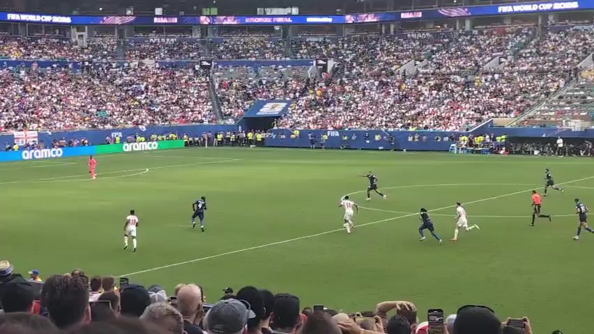 A wide shot of a soccer game in a packed stadium. The field is green, with white lines marking the boundaries. Several players in white and blue uniforms are on the field, engaged in a game. One player in a blue uniform is on the ground. Spectators fill the stands, many holding up cameras and phones. The stadium is large, with a blue and white roof structure visible. Advertising boards line the field. The atmosphere appears to be energetic and exciting.
