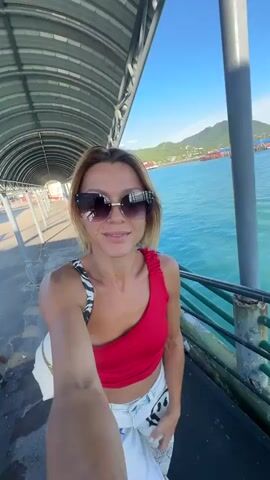 This selfie-style video features a woman with blonde hair, wearing sunglasses, a red one-shoulder top, and white pants with a graphic print. She is standing on a covered pier, with a large white ferry docked beside her. The ferry has "SEATRANS" printed on its side. The woman is smiling and appears happy. She is holding a white bag over her shoulder and takes the video in various parts of the pier.

In the background, the turquoise water of the ocean is visible, with boats and a lush, green hillside in the distance. The sky is clear and blue, indicating a sunny day. The scene suggests a tropical or coastal location, with the ferry indicating travel by sea. The time of day appears to be late morning or early afternoon due to the bright sunlight and shadows. The woman is likely on vacation or embarking on a journey.

The setting is Nathon, Thailand, a port town known for its ferry services. The presence of the ferry and the pier are the main environmental cues confirming the location. The woman's attire and joyful demeanor contribute to a feeling of leisure and adventure.