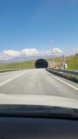 A car's perspective, looking forward towards a tunnel entrance on a sunny day. A red and white striped pole is visible to the right of the road. The car enters the tunnel, and the lighting changes to a series of yellow lights lining the tunnel ceiling. The tunnel curves to the right, and the view becomes darker and hazier.