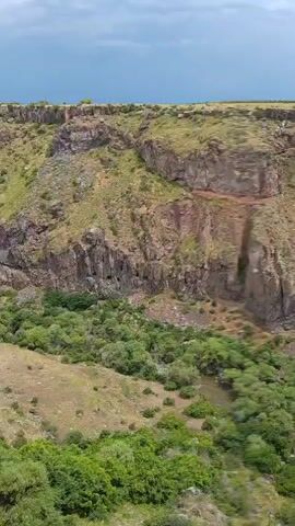 The video shows a drone perspective of a canyon. The canyon walls are steep and rocky, with sparse vegetation on the slopes. The bottom of the canyon is filled with trees and shrubs. A body of water, possibly a river or stream, is visible at the bottom of the canyon. The sky is cloudy. In the distance, a building with a red roof can be seen on top of the canyon rim. The drone appears to be flying forward and slightly downwards, offering a panoramic view of the canyon.