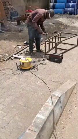 Here's a detailed description of the video:

The video shows a man working on a metal structure outdoors. He is wearing a red and black plaid shirt, dark pants, brown sandals, safety glasses, and a white covering on his head. He is using a grinder to smooth the edges of the metal frame. 

The metal frame appears to be a rectangular structure with several supports inside. A black welding machine is on the ground next to him. There is also a yellow electrical cord reel nearby.

In the background, there are other people, some sitting and watching. There are also blue barrels stacked up, along with various metal scraps and tools scattered around. The ground is paved and appears to be an outdoor workspace. The video pans to show a curb with a yellow line on the pavement.