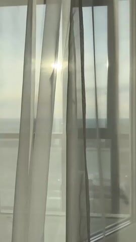 A medium, still shot captures a window covered by semi-transparent curtains. The sun is visible through the window, casting a bright glow. Outside the window, a cityscape or landscape is faintly visible. The frame of the window is white, and the curtains appear to be a light gray or white color. There are no people or actions visible in the scene.