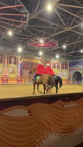 Two individuals dressed as Roman soldiers, complete with helmets and red capes, ride horses in an arena. The horses are brown and black. The arena has a dirt floor and is decorated with backdrops resembling ancient Roman architecture and murals. String lights are visible overhead. The riders appear to be performing or participating in a reenactment.