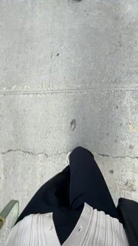 The video shows a first-person perspective of someone walking on a concrete sidewalk. The person is wearing black pants, a white cardigan, and sneakers. A green purse is visible on the left side of the frame. In the first frame, a small circular metallic object is embedded in the concrete. As the person walks, their feet and legs are the primary focus, with the background blurred. The movement is steady and appears to be on a flat surface.