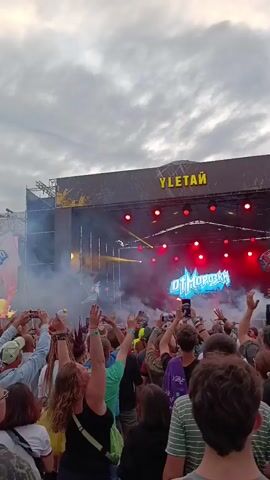 A large crowd of people are gathered outdoors at a concert. They are standing and facing a brightly lit stage where a DJ, identified by the neon sign "DJ MOROZOV," is performing. Smoke machines are in use, creating plumes of white smoke that drift across the stage and into the crowd. Many people in the audience are holding up their phones to record the performance. The sky above is cloudy and overcast. The stage has a large banner overhead with the word "УЛЕТАЙ" written on it.