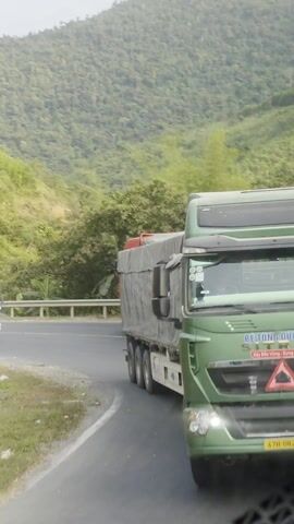 The video shows two trucks driving on a winding road through a mountainous area covered with trees. The first truck is green and has the words "XE TUNG DUC SON" on its front. The second truck is white with a silver cargo container. A red truck carrying what appears to be lumber or branches follows the white truck. The road is paved with yellow lines.