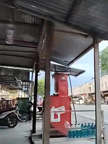 The video shows a street scene viewed from under a metal-roofed shelter. On the left is a red fuel dispenser labeled "PERTAMINE PASTI." Several bottles of water are lined up beneath the dispenser.  A street runs past in front, with two-story buildings on the opposite side. There are various businesses, including what appears to be a convenience store. Several motorcycles are parked in front of the buildings. Traffic is light, with several scooters passing by. The sky is overcast. The video seems to be shot in a Southeast Asian country based on the style of the building and scooters.