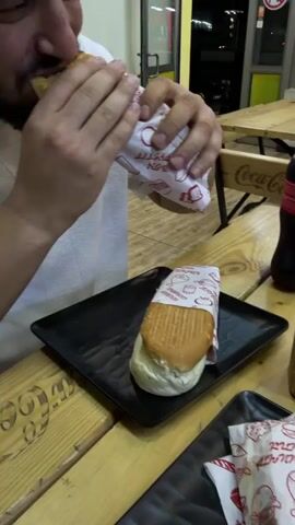 A person is shown eating a sandwich while seated at a wooden picnic table, with another wrapped sandwich on a black square plate on the table. A bottle of Coca-Cola and a small wooden box are also on the table. The sandwich is partially wrapped in white paper with red lettering and burger illustrations. The person is wearing a white shirt. The background shows a window and other tables. The person takes a bite of the sandwich.