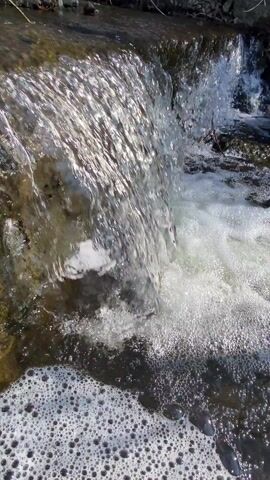 The video shows water cascading over a small, rocky waterfall. The water is clear and bubbly, with a large amount of foam accumulating at the base of the fall. The surrounding rocks are dark and wet, with some moss growth visible. The scene is well-lit, with sunlight reflecting off the flowing water.