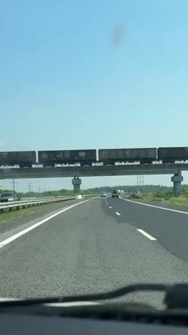 A train carrying cargo cars moves across a railway overpass above a highway. The highway has several lanes and is mostly empty, with only a few cars visible in the distance. The sky is clear and blue, indicating a sunny day. The landscape around the highway appears to be rural, with greenery and trees visible on the sides.