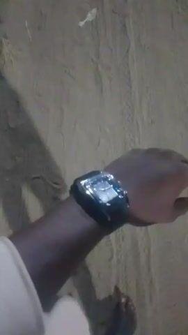 A person wearing a black watch with a silver case is walking on a sandy surface. The person is wearing a white shirt. The view is mostly focused on the wrist with the watch. The surrounding environment appears to be dimly lit.