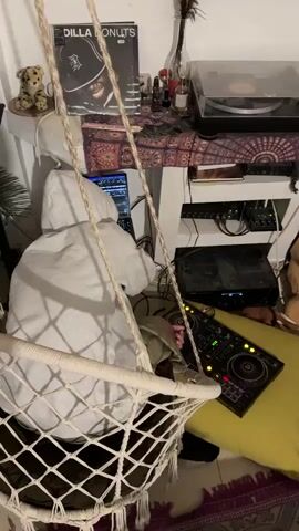 A person, seen from behind and wearing a light grey hooded sweatshirt and khaki-colored pants, is seated and actively engaged with music production equipment in an indoor setting. Their right hand is on a black DJ controller with illuminated buttons, which rests on a yellow cushion on the floor. In front of them is a laptop displaying a digital audio workstation (DAW) interface.

In the immediate foreground, a white, woven macrame-style hanging chair (swing) partially obstructs the view of the person.

The background features a white multi-tiered shelving unit acting as a music station. On the top shelf, a vinyl record album titled "DILLA DONUTS" by J Dilla is prominently displayed, showing an image of a man in a cap. Also on this shelf are a small plush toy, a potted plant, several small bottles, a dark feather in a vase, and a turntable with a record. The middle shelf is partially covered by a patterned textile and holds additional electronic audio equipment. More electronic gear and a tangle of cables are visible on the bottom shelf.

A patterned rug with red and orange tones is visible on the floor in the bottom right corner. The lighting appears to be indoor, possibly artificial. The audio component includes music with a clear beat and Arabic vocals, indicating active music mixing or production. The scene depicts a focused individual in a personalized creative or home studio space. The specific location of Mitzpe Ramon, Israel, is not visually discernable from the indoor environment.