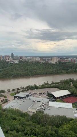 This is a high-angle, vertical video showcasing an aerial view of a city and its surrounding natural landscape. The primary subject is the expansive cityscape in the background, characterized by a variety of buildings, including tall high-rises and lower-rise structures, interspersed with dense green trees.

In the mid-ground, a wide, muddy river flows horizontally across the frame, separating the city from a strip of lush forest. Along the near bank of the river, there are several large, modern buildings with grey roofs, appearing to be industrial or recreational facilities. Adjacent to these buildings, a parking lot filled with vehicles is visible. Further to the right, there are two red tennis courts and a small playground area with colorful equipment.

The foreground is dominated by a dense canopy of green trees, suggesting the viewpoint is from a height, possibly a tall building or a natural vantage point like a cliff.

The weather appears overcast with dramatic cloud formations in the sky, hinting at potentially gloomy or stormy conditions. The time of day is ambiguous due to the cloud cover, but it seems to be daytime. No people are distinctly visible in the frame.

The overall scene depicts an urban environment integrated with significant natural elements, highlighting the proximity of the city to the river and its forested banks. There is no visible text in the media file.