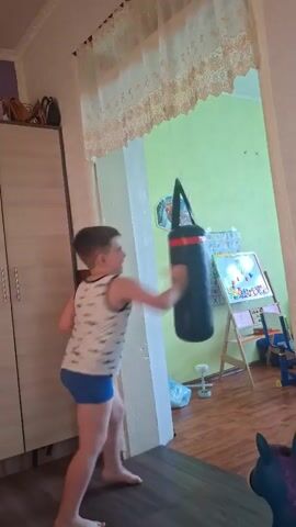 A young boy in a white tank top with a pattern and blue shorts is practicing kicks and punches on a black punching bag that hangs from the ceiling. He is standing in a room with a wooden floor. To the left of the boy is a tall, light-colored cabinet with two doors. To the right, a light green wall is visible, adorned with a framed picture of a cartoon character, a small basketball hoop, and a drawing of a Christmas tree. A tripod easel with a drawing on it stands in front of the green wall, with a toy blue chair in the lower right corner of the frame. A patterned curtain hangs across the top of the doorway. The boy moves his arms and legs in a fluid motion, engaging with the punching bag.