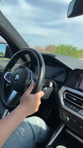 The video captures the interior of a car from the driver's perspective. A person's right hand is visible on the steering wheel, which features a BMW logo. The driver's leg, clad in light-colored jeans, is also partially visible. The dashboard and a modern infotainment system dominate the center console.

The infotainment screen displays "Media/Radio" and "No device connected.", along with "Communication" and "Add new device.". The time shown is 09:42, and the internal temperature is 24 degrees Celsius. Through the windshield, a bright sky with scattered clouds is visible, along with an open road and a distant landscape of fields and trees, indicative of driving through a rural or semi-rural area. The scene takes place in Voyvodovo, Bulgaria. The sound of a vehicle in motion can be heard.