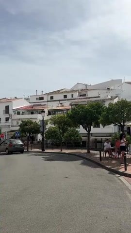 The video shows a view of a street scene with buildings and people. In the foreground, there is a paved street curving to the right, with a sidewalk made of reddish and tan bricks. A white planter with green plants sits on the right side of the sidewalk. Several trees with green foliage are planted along the sidewalk. In the middle ground, there is a row of white buildings with red-tiled roofs, typical of Mediterranean architecture. Some people are seated at tables outside what appears to be a cafe, with umbrellas providing shade. One person is walking along the sidewalk. Further back, more buildings and trees are visible, with some structures that appear to be cell towers on a hill. The sky is partly cloudy. The overall impression is a sunny day in a town with a pleasant outdoor atmosphere.