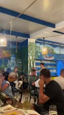 The video shows an open-air restaurant with live music. A musician with a guitar and a singer are performing for a crowd of seated diners. The restaurant's interior has a Mediterranean theme with white and blue color schemes. Murals of Santorini-like scenery adorn the walls. People are seated at tables enjoying their meals and drinks, some appear to be listening to the music. Outside the restaurant, street lights and passing pedestrians are visible. The camera moves around, focusing on different tables of diners and the musicians performing.