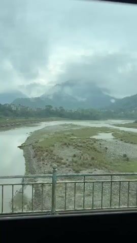 The scene is a cloudy day view of a wide, shallow river meandering through a valley. Mountains are visible in the background, partially obscured by low-hanging clouds. The view is framed by a metal railing, likely from a bridge. Some buildings can be seen on the hillside in the distance. The river's banks are mostly gravel and sparse vegetation.