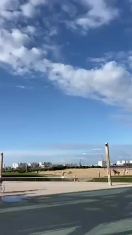 The video depicts two people riding bicycles on a paved path. They are riding away from the camera, moving from left to right, and toward a sandy area with a few buildings in the background. In the background, there is also a person on horseback in the distance. The sky is blue with scattered white clouds, and the lighting is bright. There are a few puddles on the path. There are poles on either side of the path.