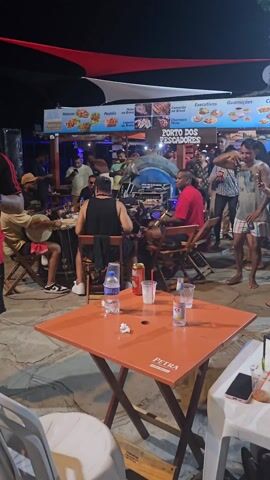 The video shows a live band playing outdoors at night in a lively setting, likely a restaurant or bar called "Porto dos Pescadores".  Four men are seated and playing instruments: one is playing a tambourine-like instrument called a pandeiro, another is playing a guitar, a third plays a percussion instrument, and the fourth sits behind a DJ mixer. Several people are standing around, some dancing and socializing. Tables with drinks and bottles are visible in the foreground.  The area is lit with string lights and under the roof of an open-air structure.  The name "PETRA" is printed on the side of a table.  The atmosphere appears relaxed and festive.