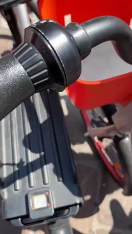 The video shows the front of several bicycles, including handlebars with QR codes and orange baskets. The bicycles are red, white, and black. People are standing around the bicycles. One person wears a white t-shirt and black shorts, while another wears blue shorts. The bicycles appear to be part of a bike-sharing program. The setting seems to be a public square with buildings visible in the background.