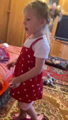A young girl with blonde hair tied in a bun with a pink bow is wearing a white t-shirt and a red dress with white hearts. She is standing in a room with a patterned rug on the floor and wooden furniture in the background. The girl is holding a toy tiara and adjusting it on her head. There is a stuffed animal on the floor near the furniture.